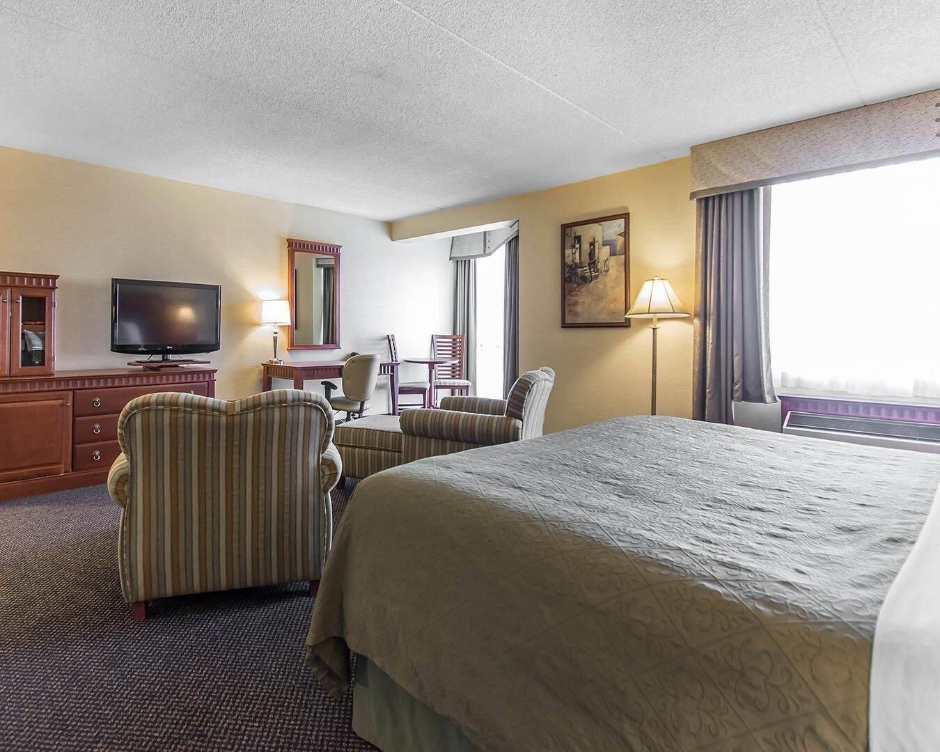 A hotel room with a large bed, a desk with a television, a chair, a lamp, and a window with curtains.