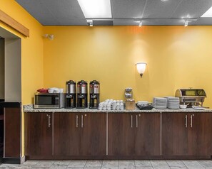Un espace pour le petit-déjeuner à l’hôtel, avec un mur jaune, un coin café équipé de quatre machines à café, un four à micro-ondes et un buffet avec des assiettes et des tasses.