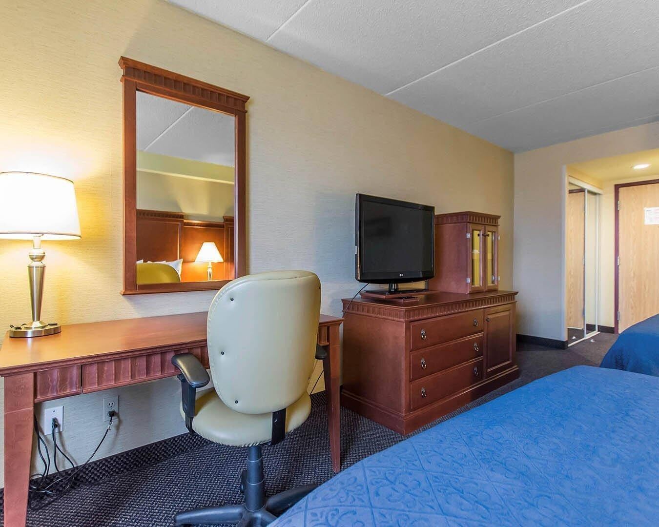 A hotel room with a bed, a desk with a lamp, a chair, a television on a wooden cabinet, and a mirror on the wall.
