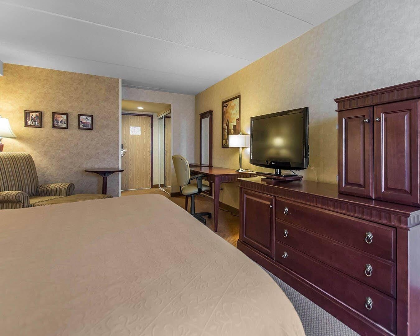 Ein Hotelzimmer mit einem großen Bett, einem Fernseher, einem Schreibtisch und einem Sessel. Eine Tür führt in einen weiteren Raum.