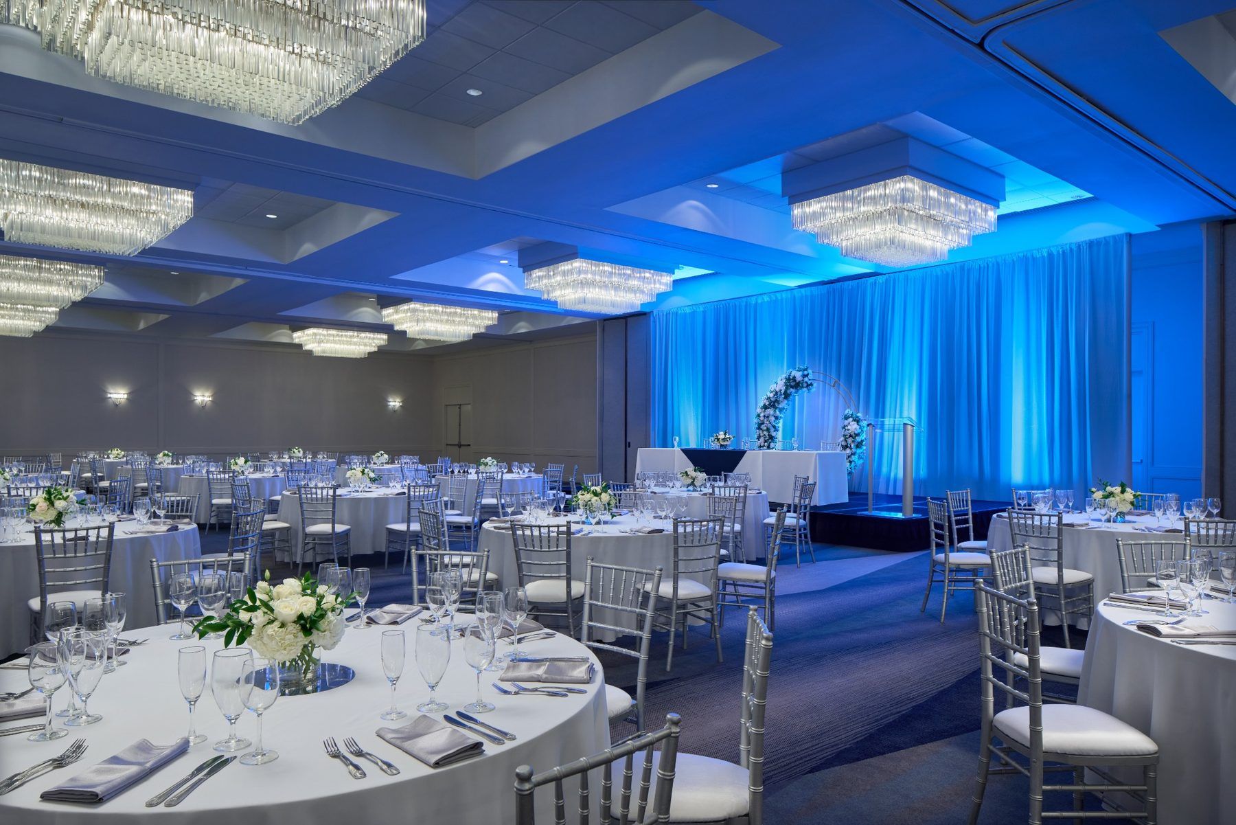 A banquet hall with round tables, chairs, and a stage with a backdrop.