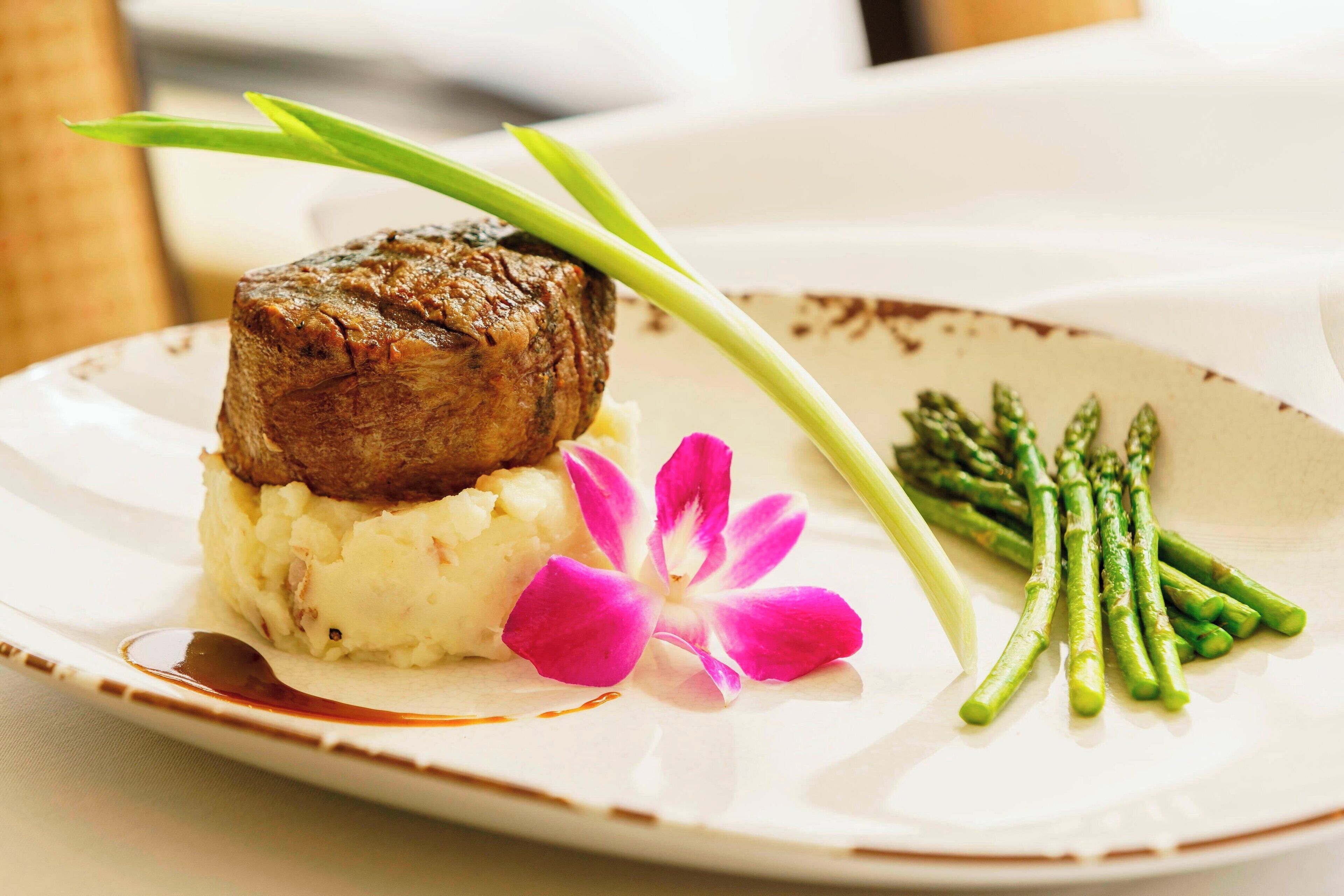Un plato con puré de papas, un trozo de carne, espárragos y una flor rosa.