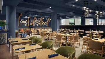 A modern restaurant interior with multiple dining tables and chairs, a bar area, and decorative wall art.