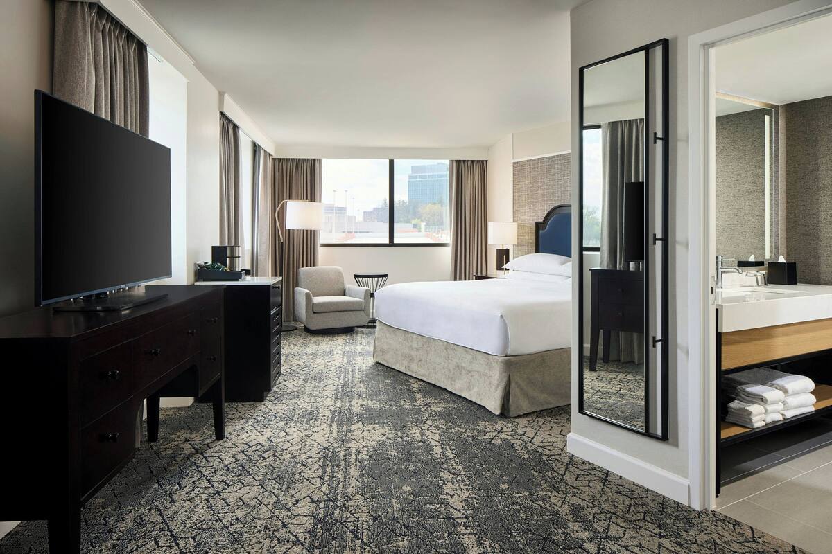 Quarto de hotel com uma cama grande, uma televisão sobre uma cômoda, uma cadeira, uma escrivaninha e um banheiro com espelho e pia.