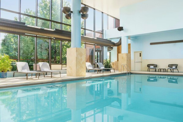 An indoor swimming pool with clear glass walls, lounge chairs, and a view of trees outside. - Sheraton Baltimore North Hotel (Towson)