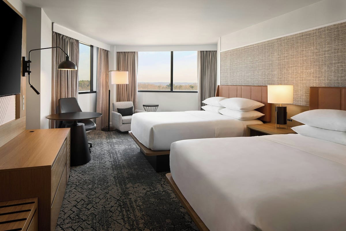 Quarto de hotel com duas camas, uma escrivaninha, uma cadeira, uma mesinha e vista para o exterior.