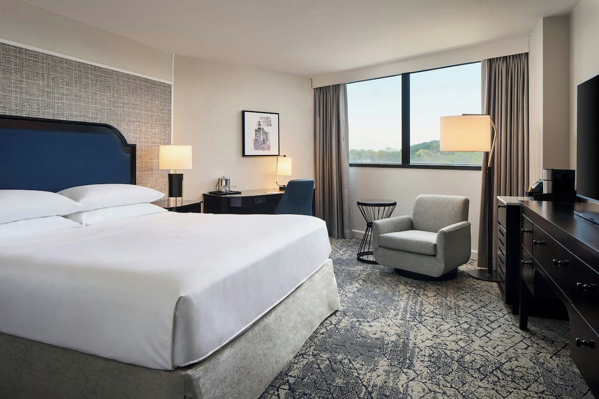 Quarto de hotel com uma cama grande, uma escrivaninha com abajur, uma cadeira, televisão e janela com vista.