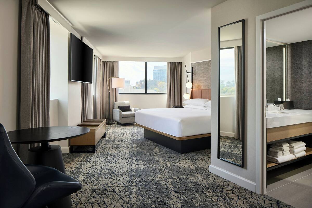 Quarto de hotel com cama, escrivaninha, cadeira, televisão, banheiro com espelho e vista da cidade.
