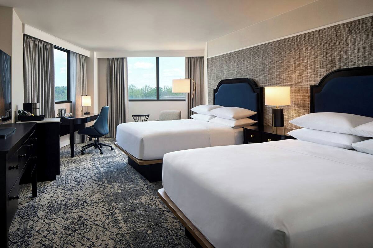 Quarto de hotel com duas camas, uma escrivaninha, uma cadeira e vista para o exterior.