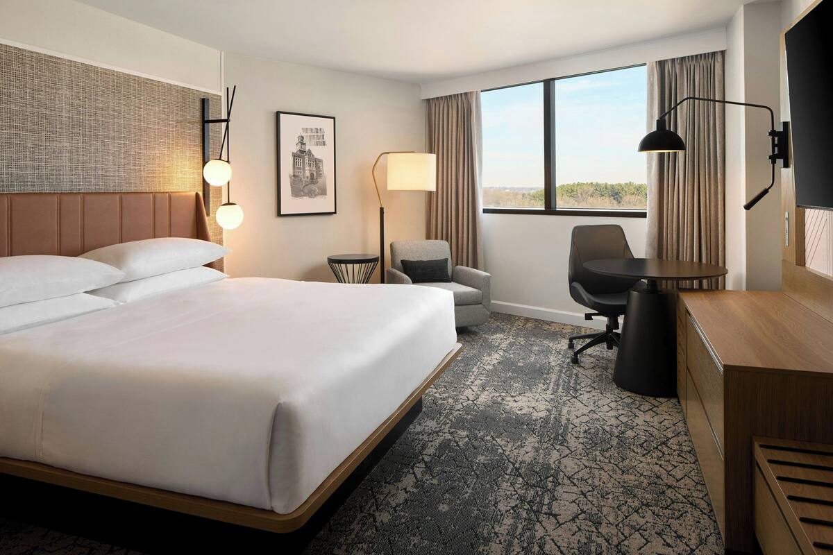 Quarto de hotel com uma cama grande, uma escrivaninha, uma cadeira, um abajur e vista para o exterior.