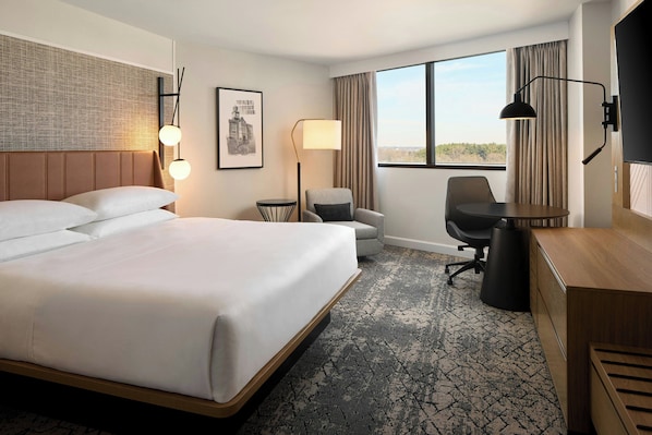 A hotel room with a large bed, a desk, a chair, a lamp, and a view of the outdoors.