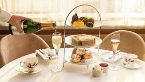 A table set for a tea service with a tiered dessert stand, a bottle of champagne being poured, and tea cups and saucers. - The Cavendish London (London)