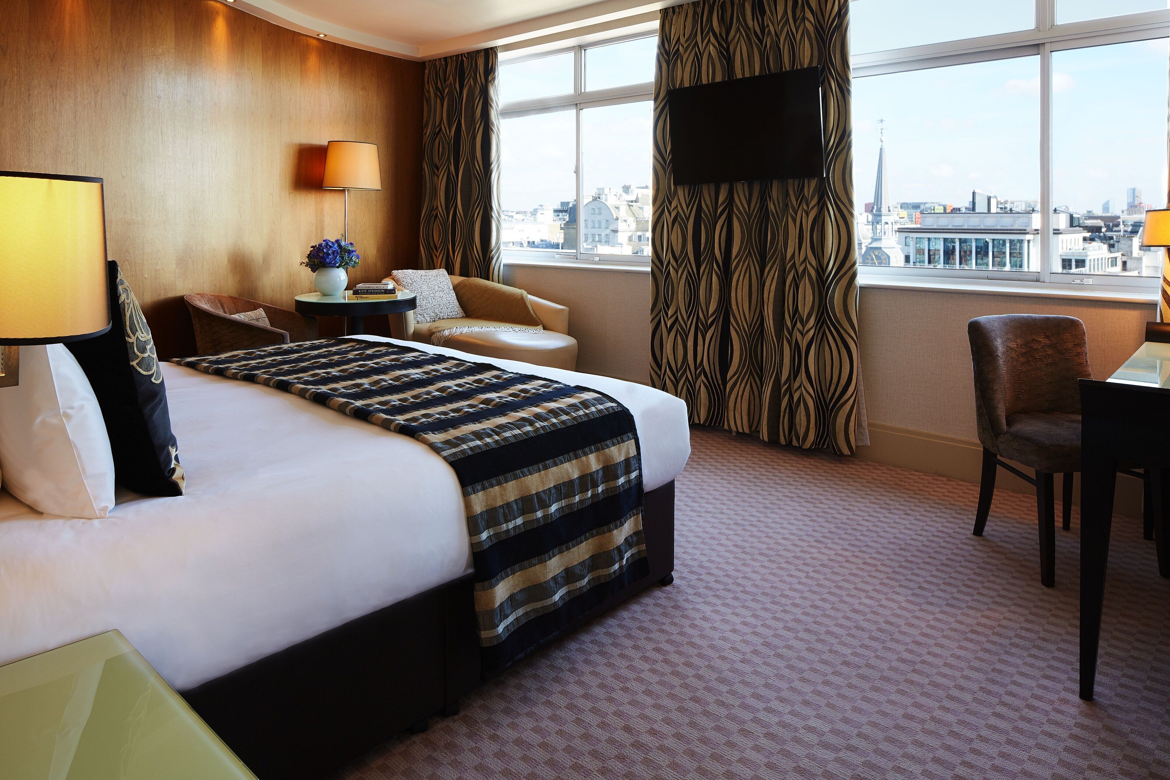 Habitación de hotel con una cama grande, un escritorio, una silla, un televisor y vistas a la ciudad desde la ventana.