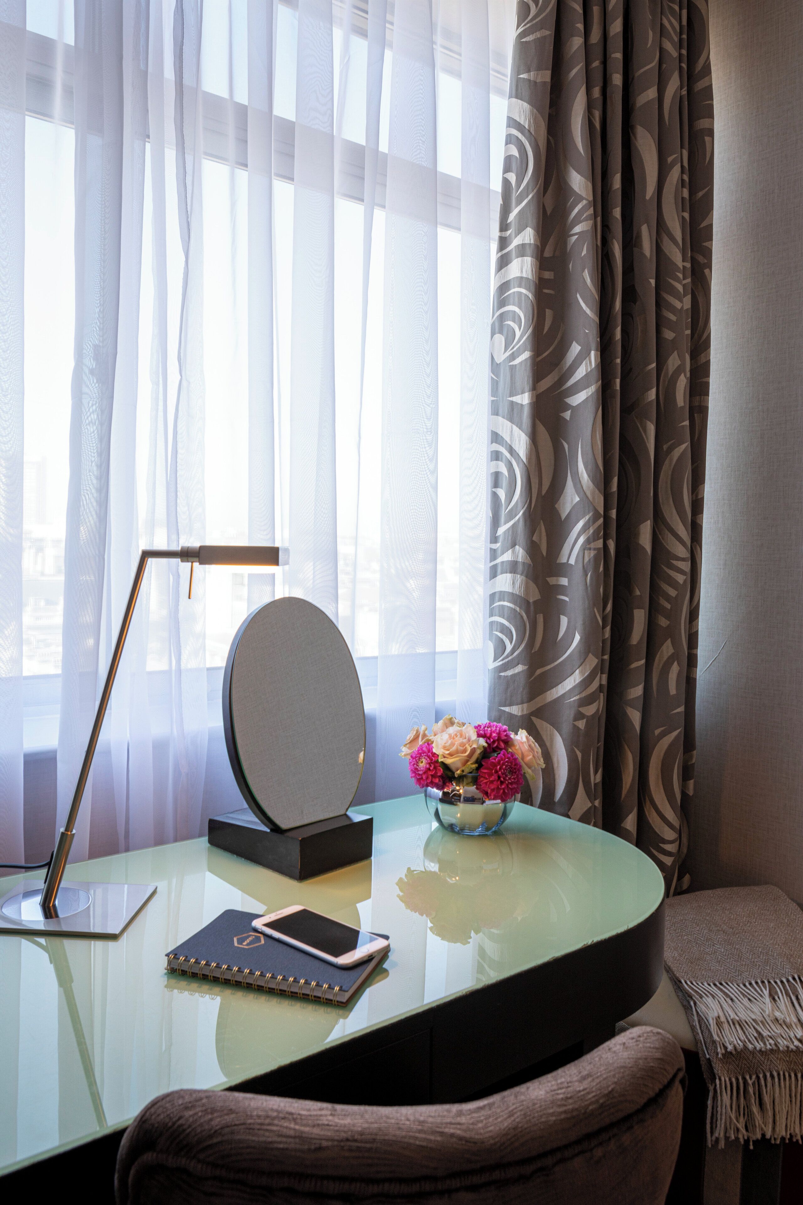 Een moderne kamer met een glazen bureau, een laptop, een smartphone, een notitieboek, een spiegel, een vaas met bloemen, een lamp en uitzicht door dunne gordijnen.