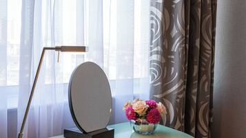 Een moderne kamer met een glazen bureau, een laptop, een smartphone, een notitieboek, een spiegel, een vaas met bloemen, een lamp en uitzicht door dunne gordijnen.