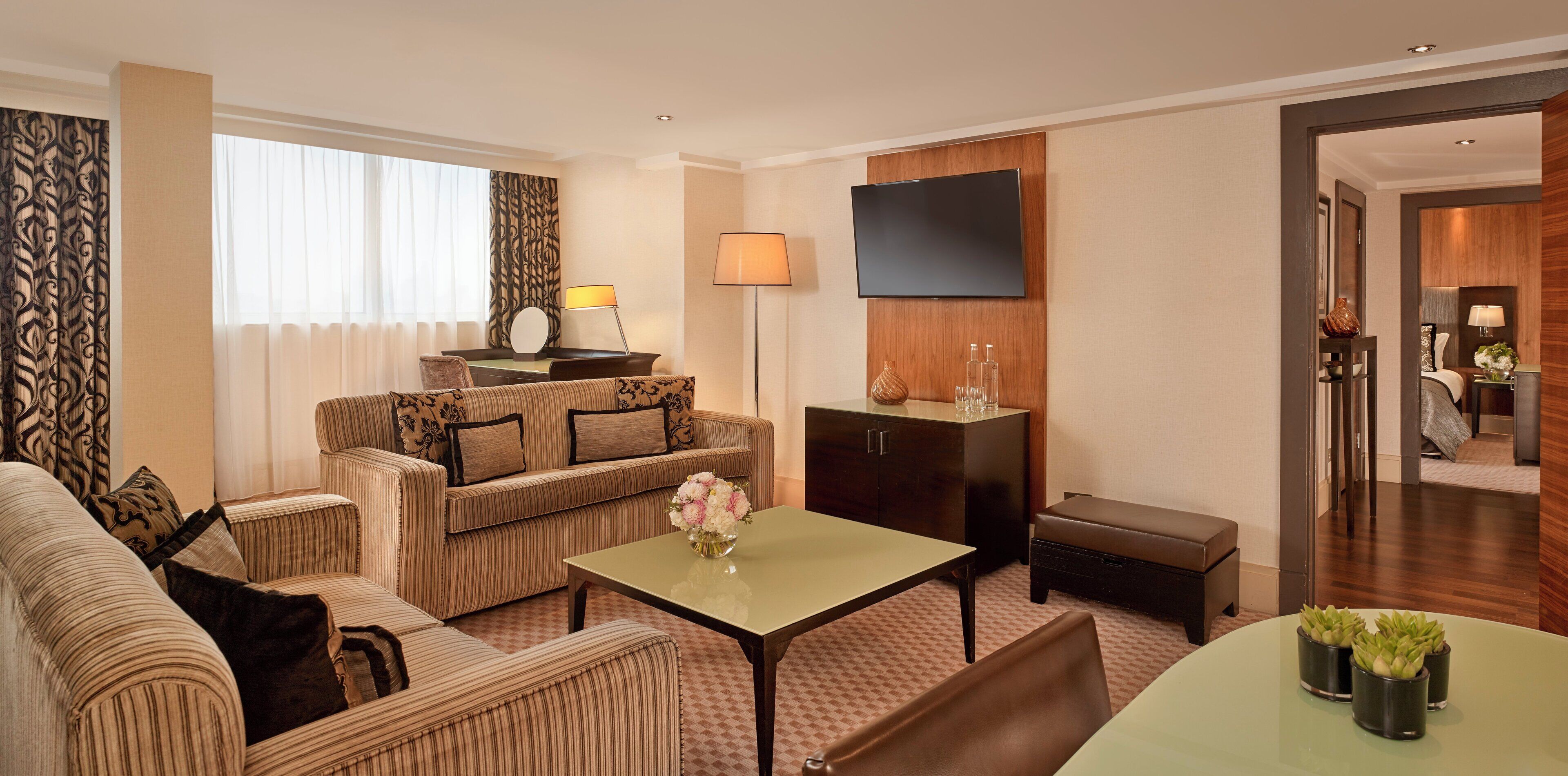 A hotel room with a sofa set, a small table with a vase of flowers, a cabinet, and a television mounted on the wall.