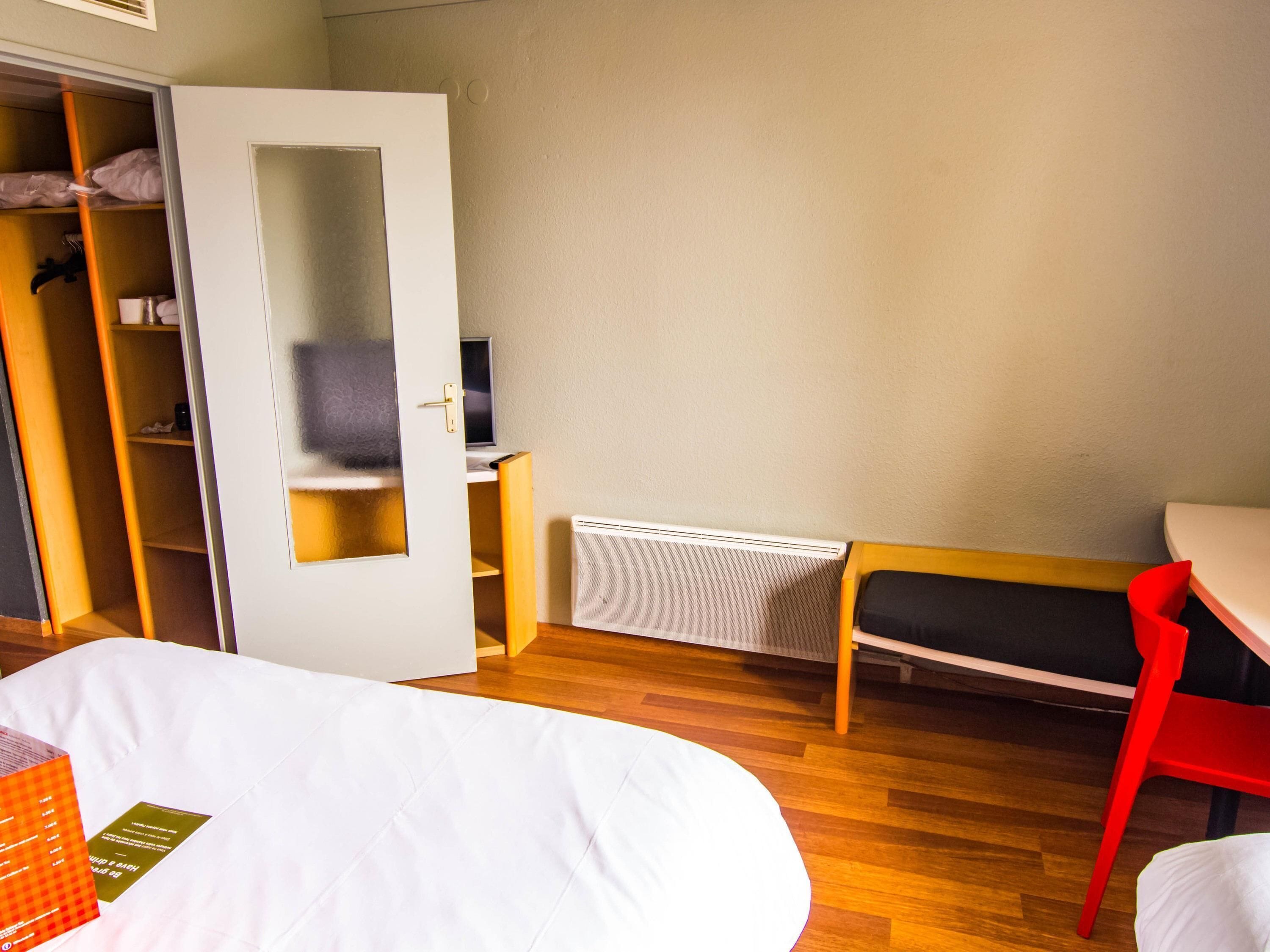 Una habitación de hotel con cama, escritorio, silla, armario y radiador.