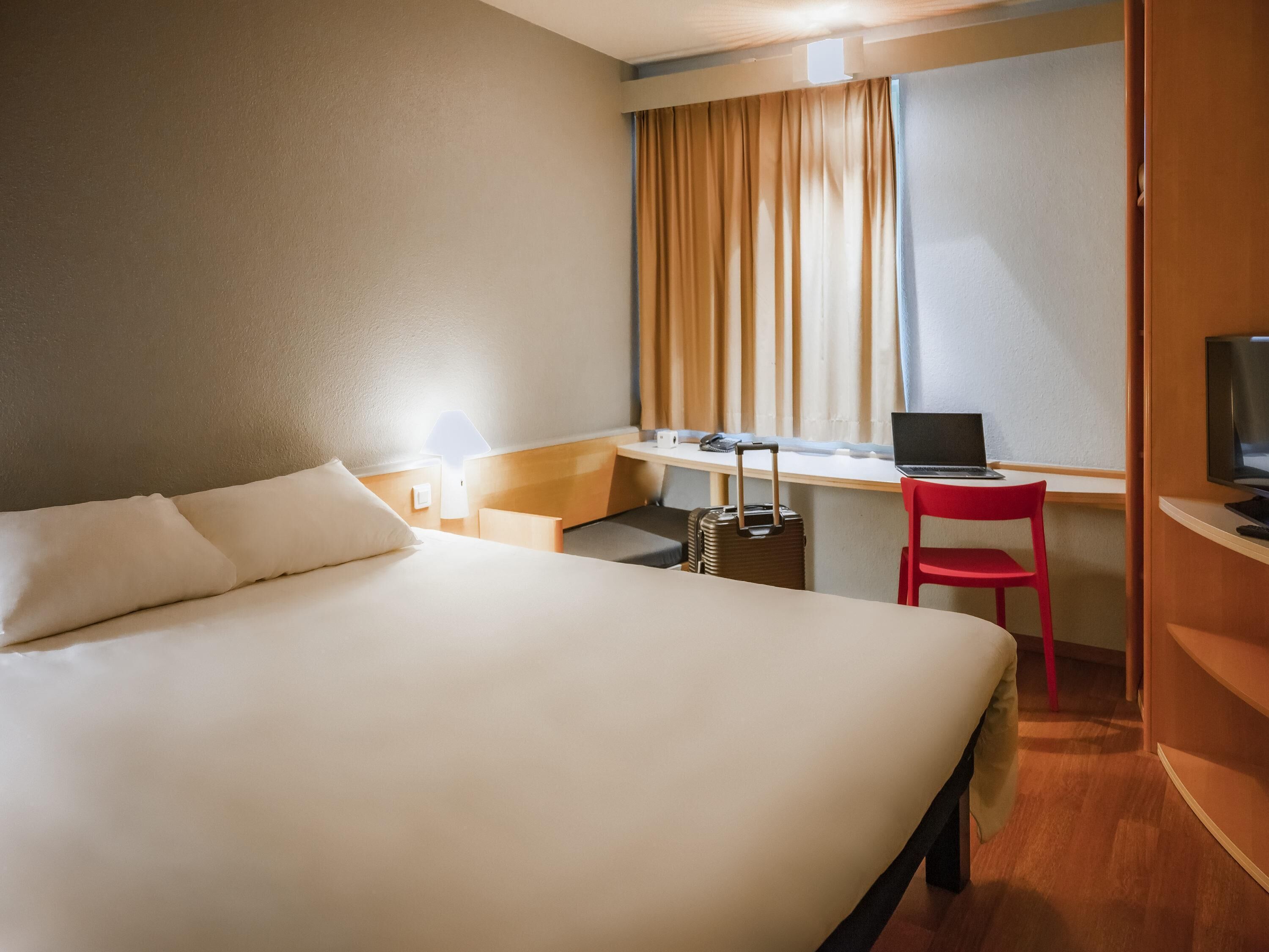 A hotel room with a bed, a desk with a laptop, a chair, a suitcase, and a TV.