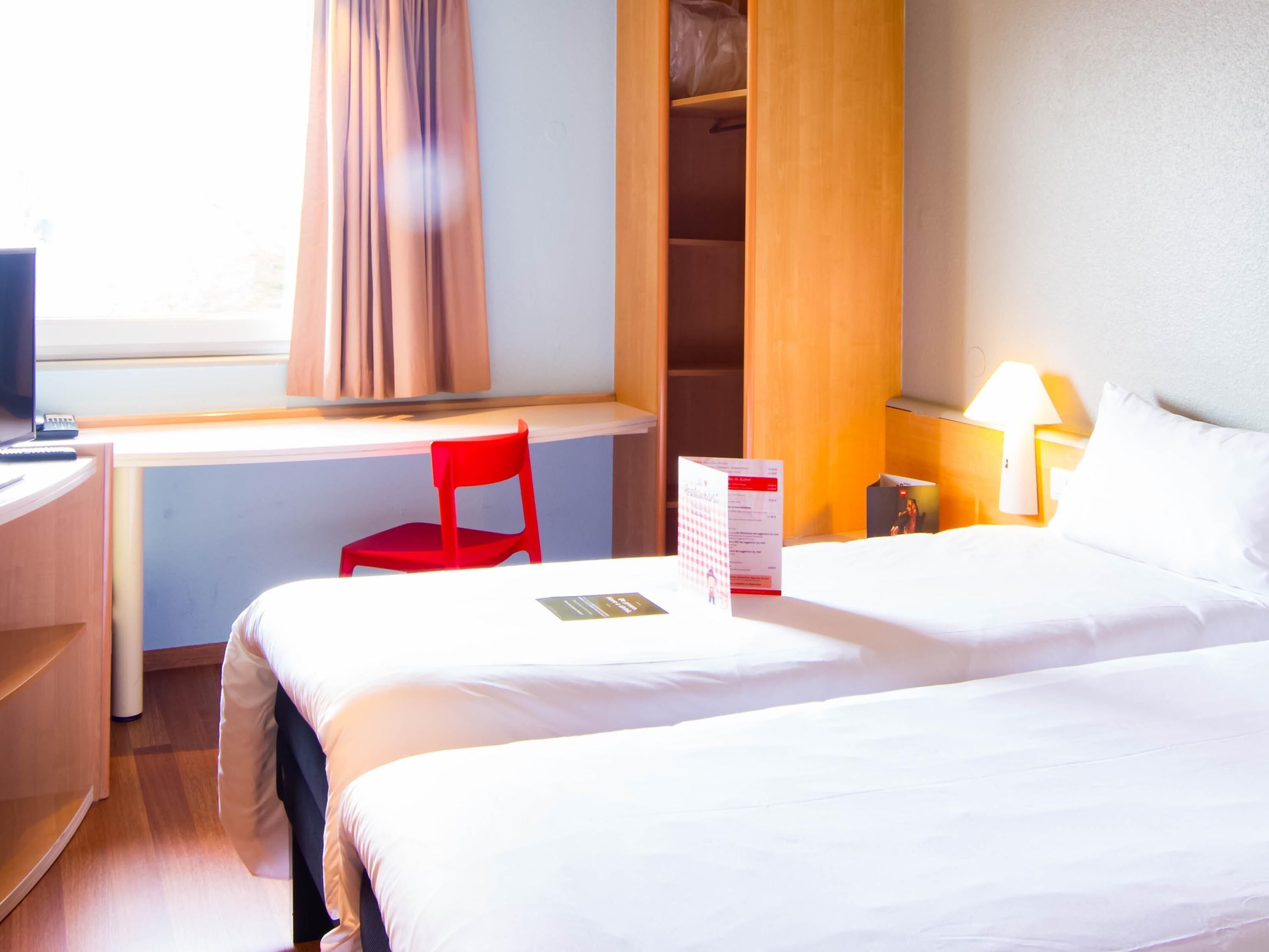 Une chambre d’hôtel avec un lit, un bureau, une chaise, une table de chevet et une fenêtre avec des rideaux.