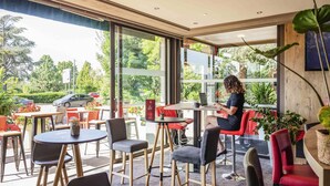 A modern cafe with large windows, outdoor seating, and a person working on a laptop. - ibis Colmar Est (Horbourg-Wihr)