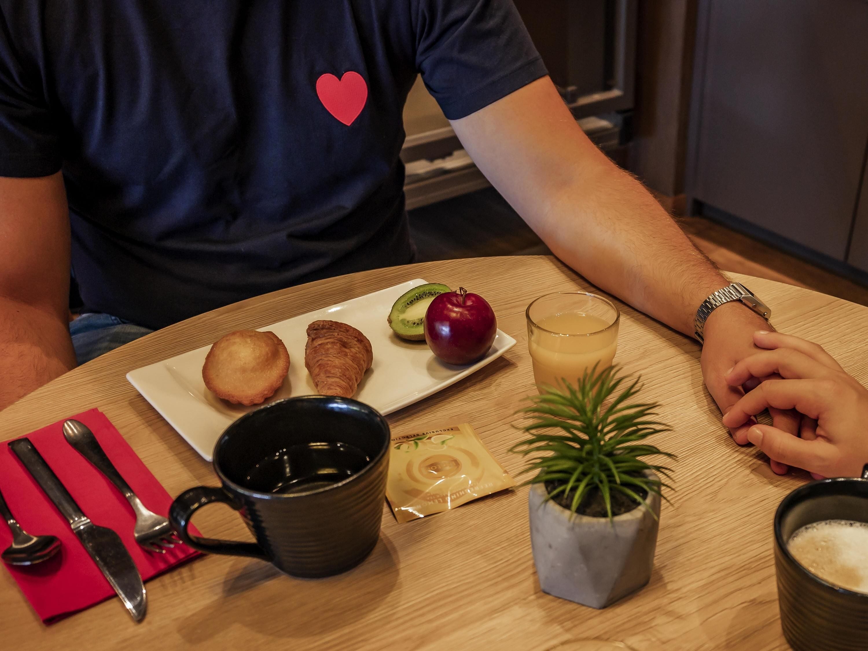 Ein Esstisch mit einem Teller Essen, einem Getränkglas, einer Tasse, Besteck, einer Pflanze und einer herzförmigen Verzierung auf einem Hemd.