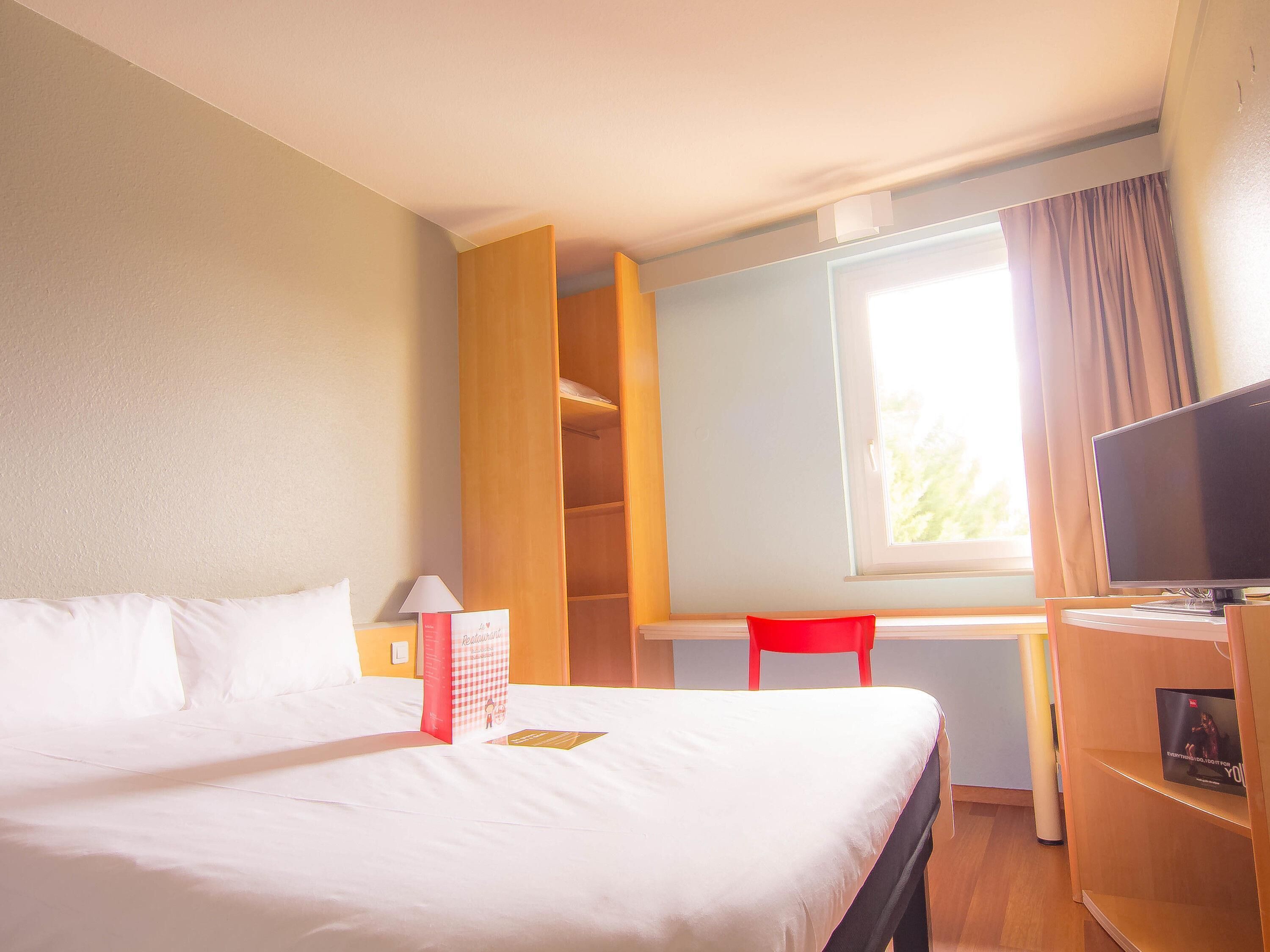 Una habitación de hotel con cama, escritorio con computadora, silla, ventana con cortinas y televisor.