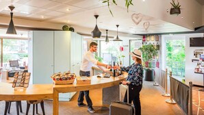 A reception desk with a man and a woman, a basket of cookies, and a suitcase. - ibis Colmar Est (Horbourg-Wihr)