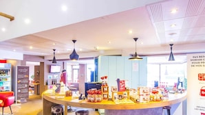 A modern interior with a central round table displaying various products, a seating area with stools, and a visible counter with a menu. - ibis Colmar Est (Horbourg-Wihr)