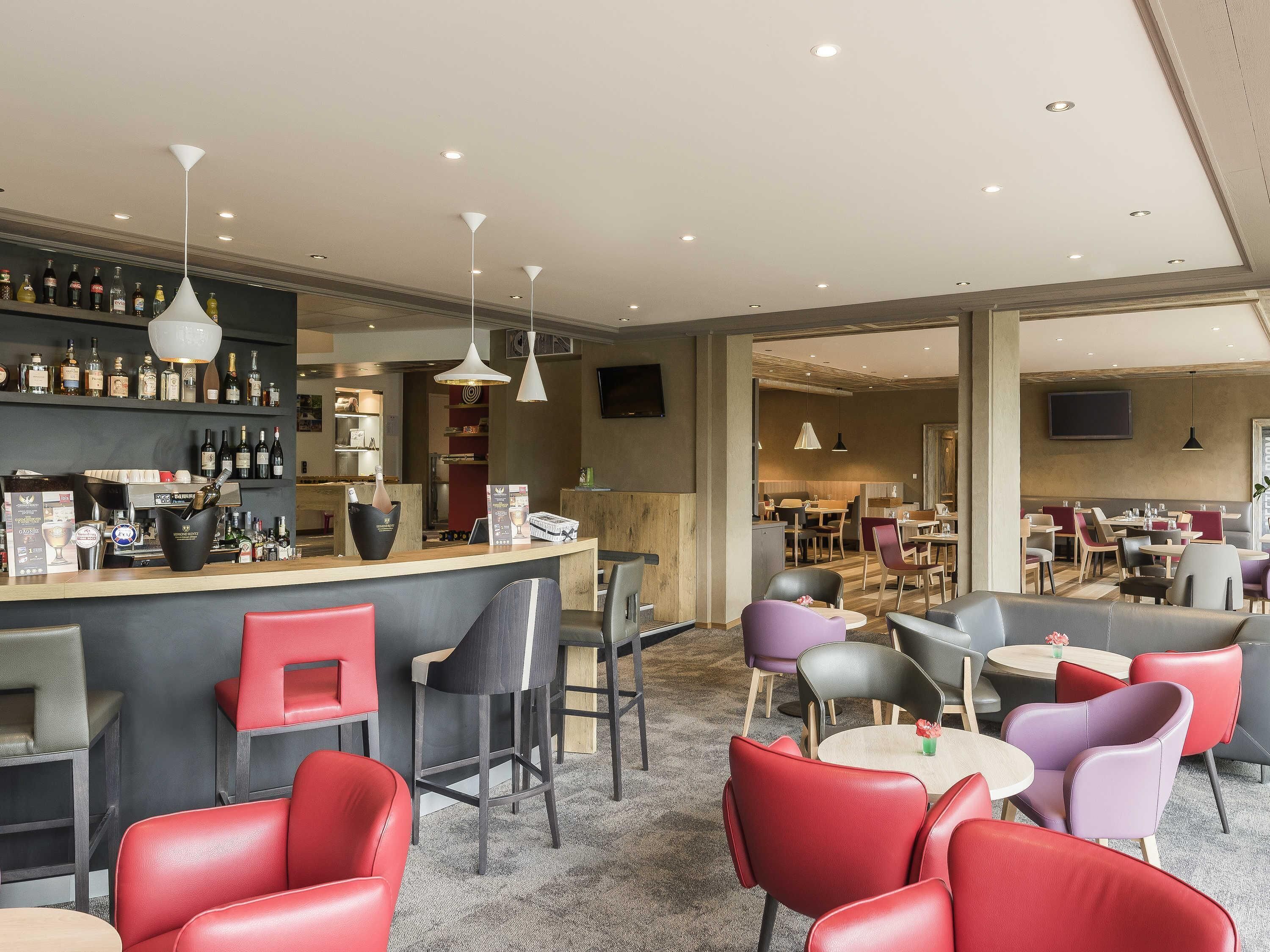 A modern bar area with a variety of seating options including red and purple chairs, a counter with a wine bottle display, and overhead lighting.