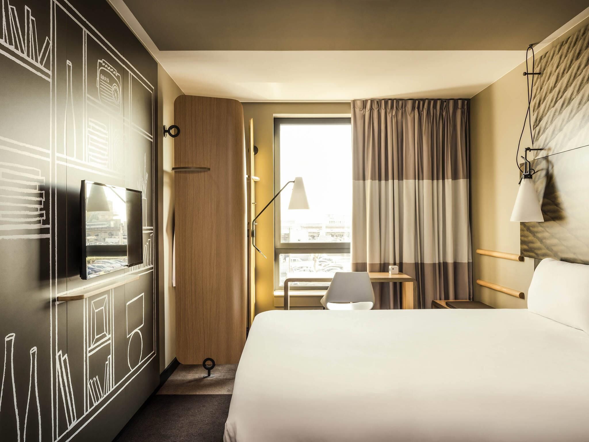 Une chambre d’hôtel comprenant un lit, un bureau avec une chaise, un téléviseur fixé au mur, une fenêtre avec des rideaux et une vue sur le paysage urbain.