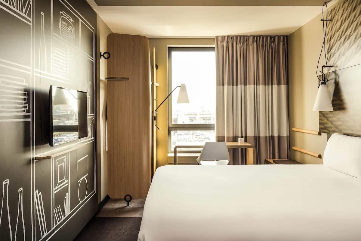 Hotelkamer met een bed, een bureau met stoel, een aan de muur bevestigde televisie, een raam met gordijnen en uitzicht op de stad.