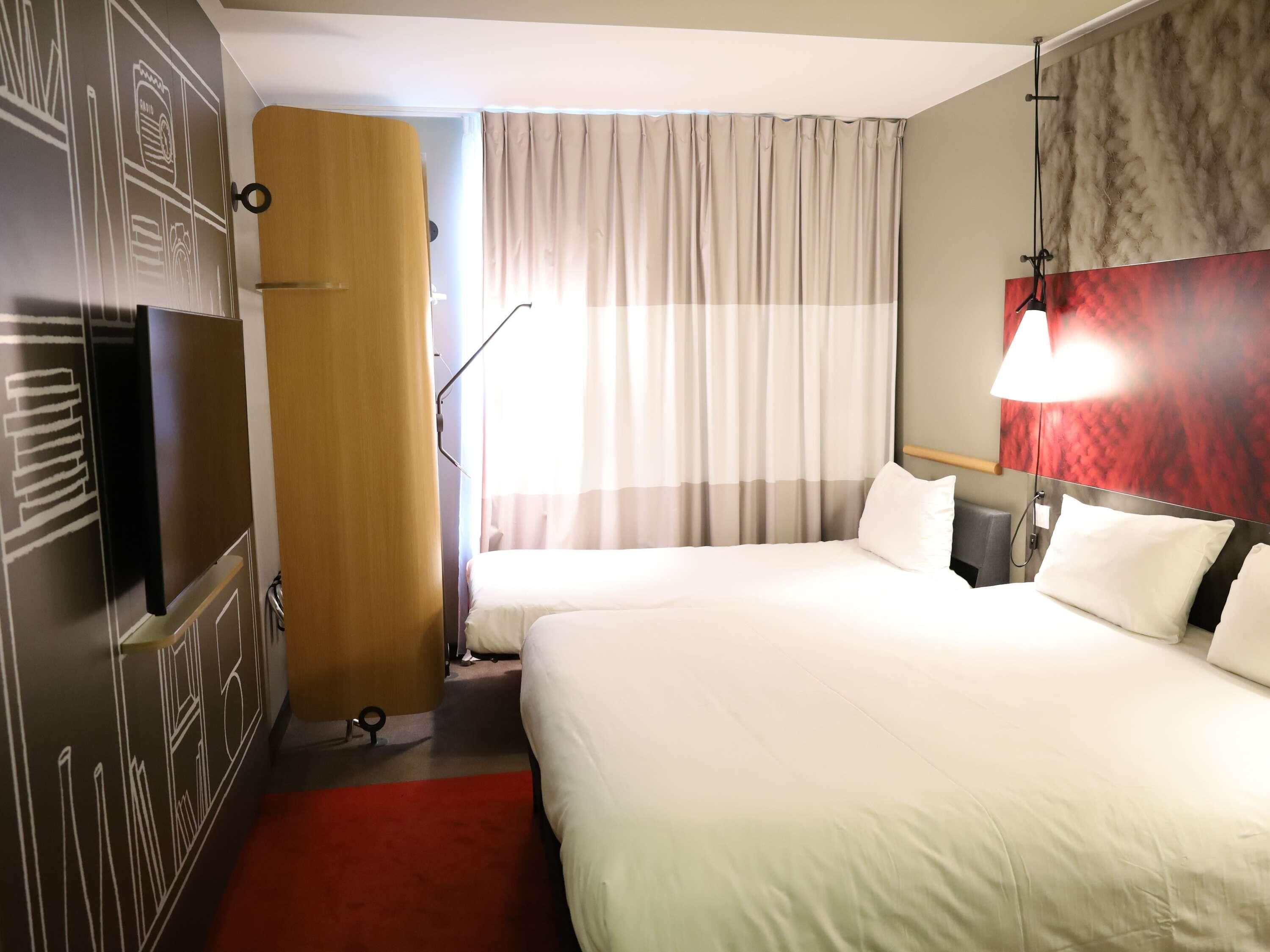 A hotel room with two beds, a red carpet, a wooden door, a large window with curtains, and a wall with a red and grey pattern.