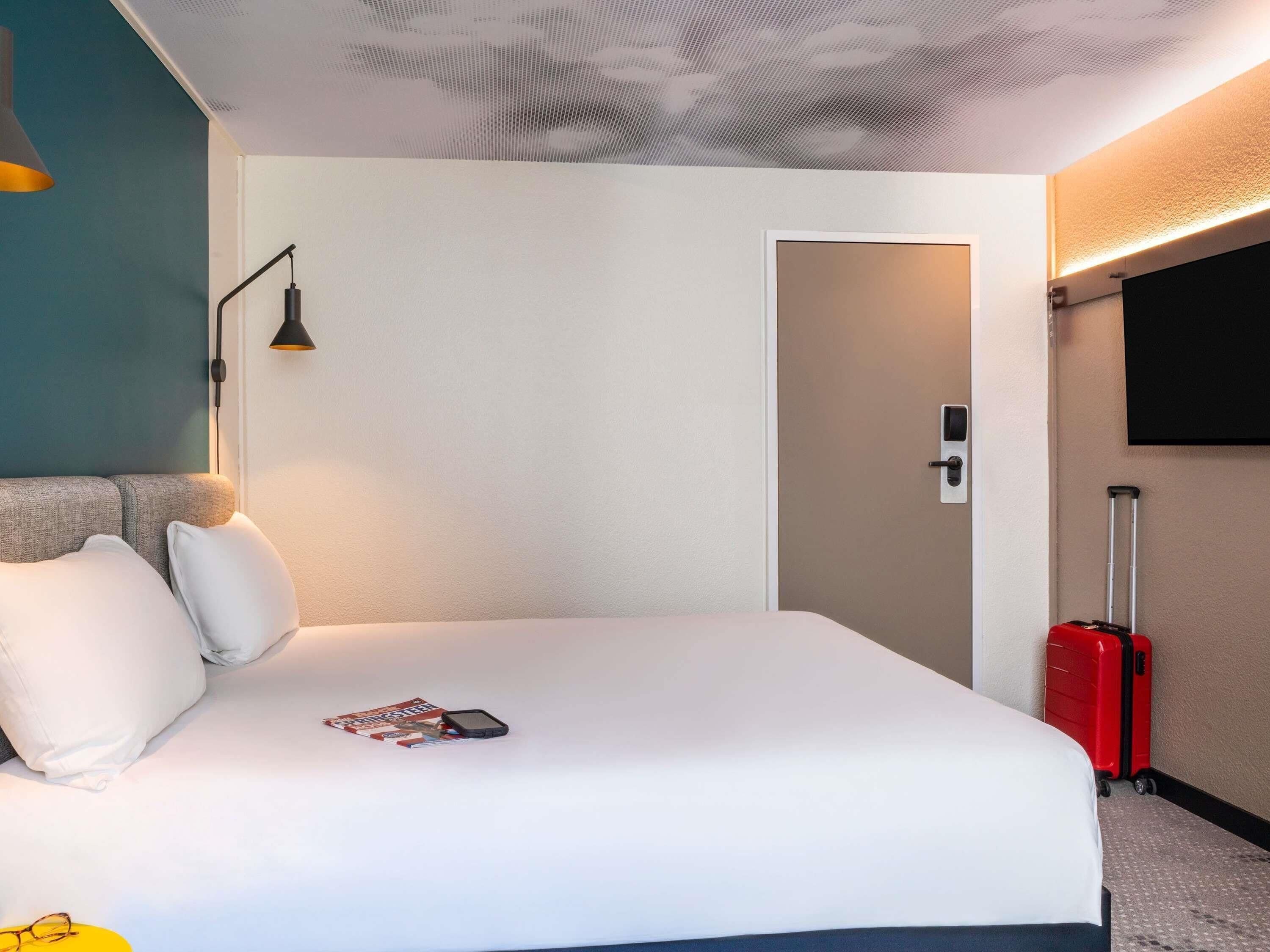 A hotel room with a bed, a TV mounted on the wall, a door, and a red suitcase.