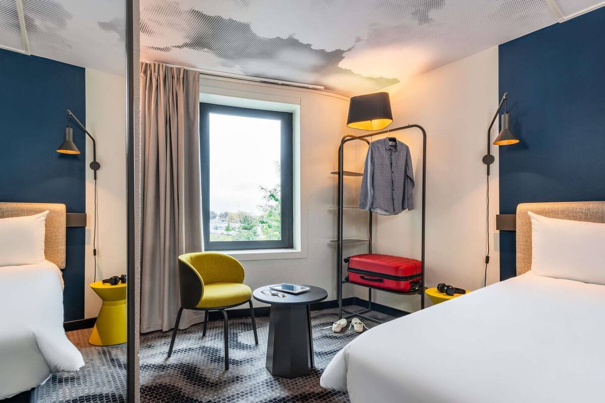 Een hotelkamer met twee bedden, een raam met uitzicht, een stoel, een tafeltje, een lamp, een kapstok met een jas en een koffer.