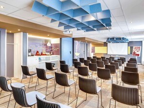 Ein Konferenzraum mit Reihen von Stühlen, einer Projektionsleinwand und einem Empfangsbereich mit Theke und Dekoration.