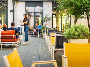 An outdoor seating area with tables and chairs, a woman standing with a tablet, and people dining in the background. - ibis Paris Coeur d'Orly Airport (Orly)