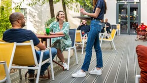 Un espace extérieur aménagé avec des tables et des chaises, une personne debout et un bâtiment en arrière-plan.