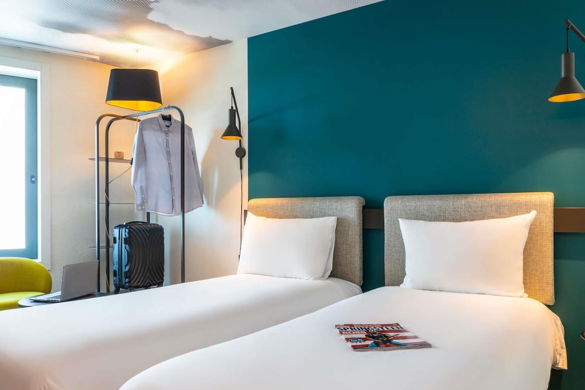 Une chambre d’hôtel avec deux lits, un bureau avec un ordinateur portable, une valise, une chaise et un magazine sur le lit.