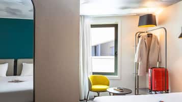 Habitación de hotel con cama, una silla, una mesita, una lámpara, perchero y maleta.
