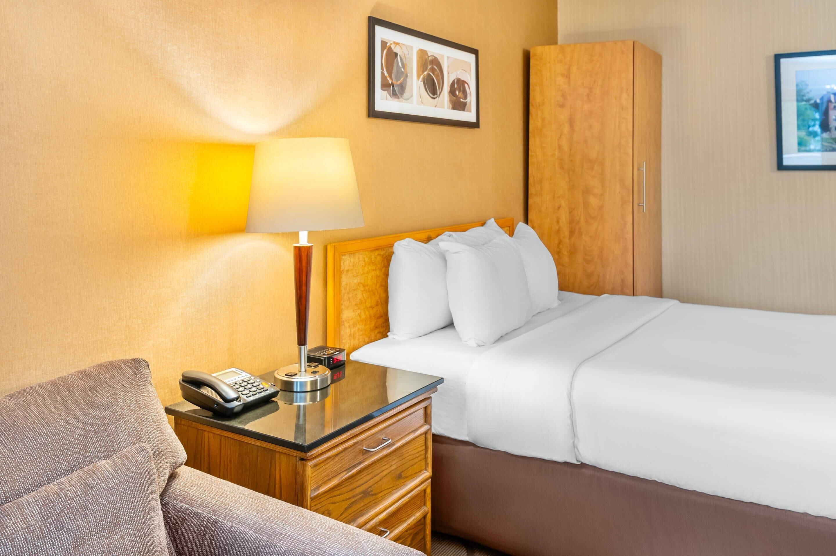 Quarto de hotel com cama, mesa de cabeceira com abajur, telefone e guarda-roupa de madeira.