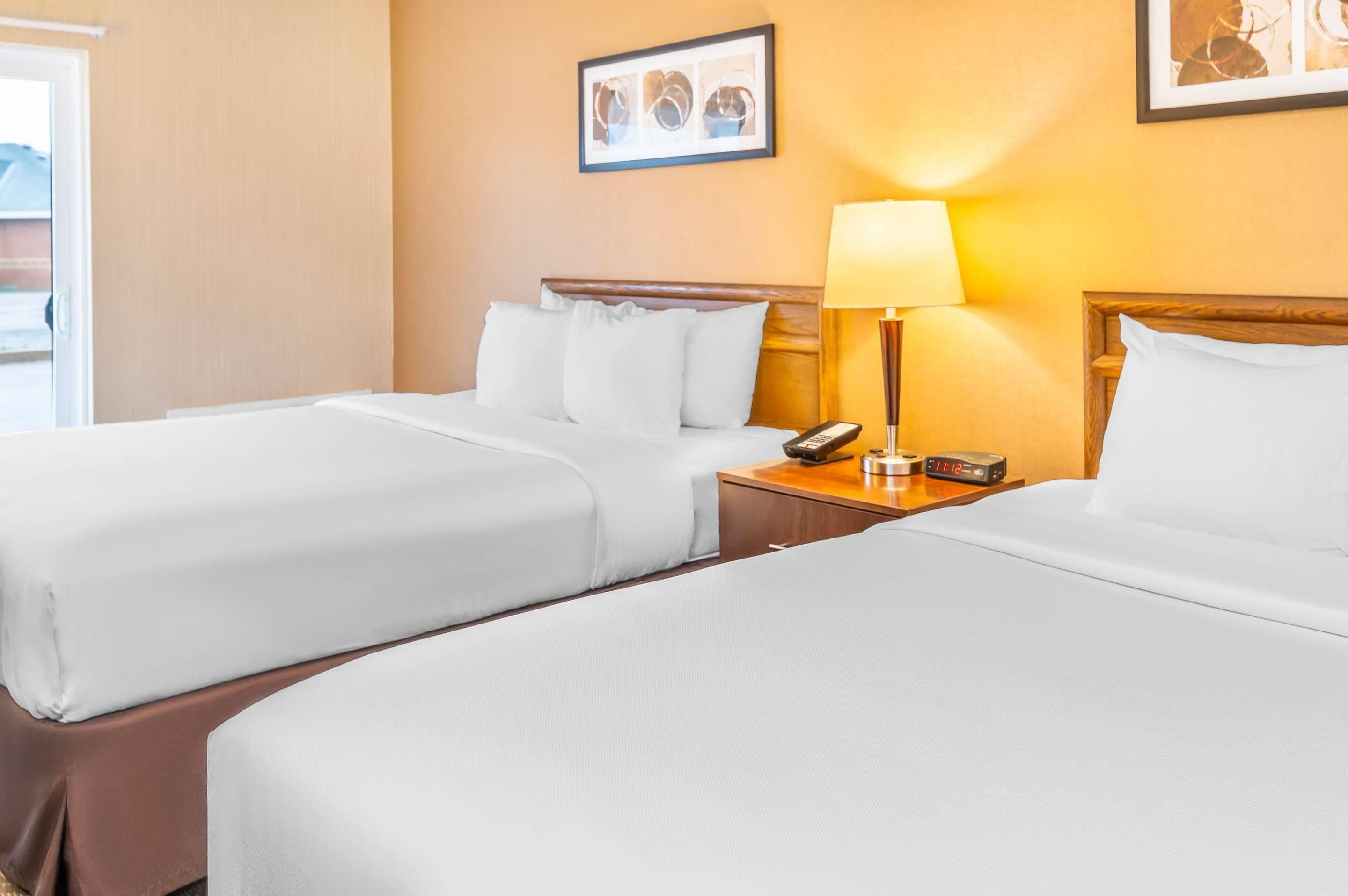 A hotel room with two beds, a nightstand with a lamp, a clock, and a framed picture on the wall.