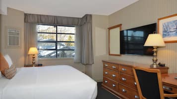 Ett hotellrum med en säng, en byrå, ett skrivbord, en lampa, en tv och ett fönster med gardiner.