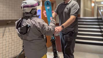 A person in a helmet and ski gear holding a pair of skis, standing on a staircase in a building.