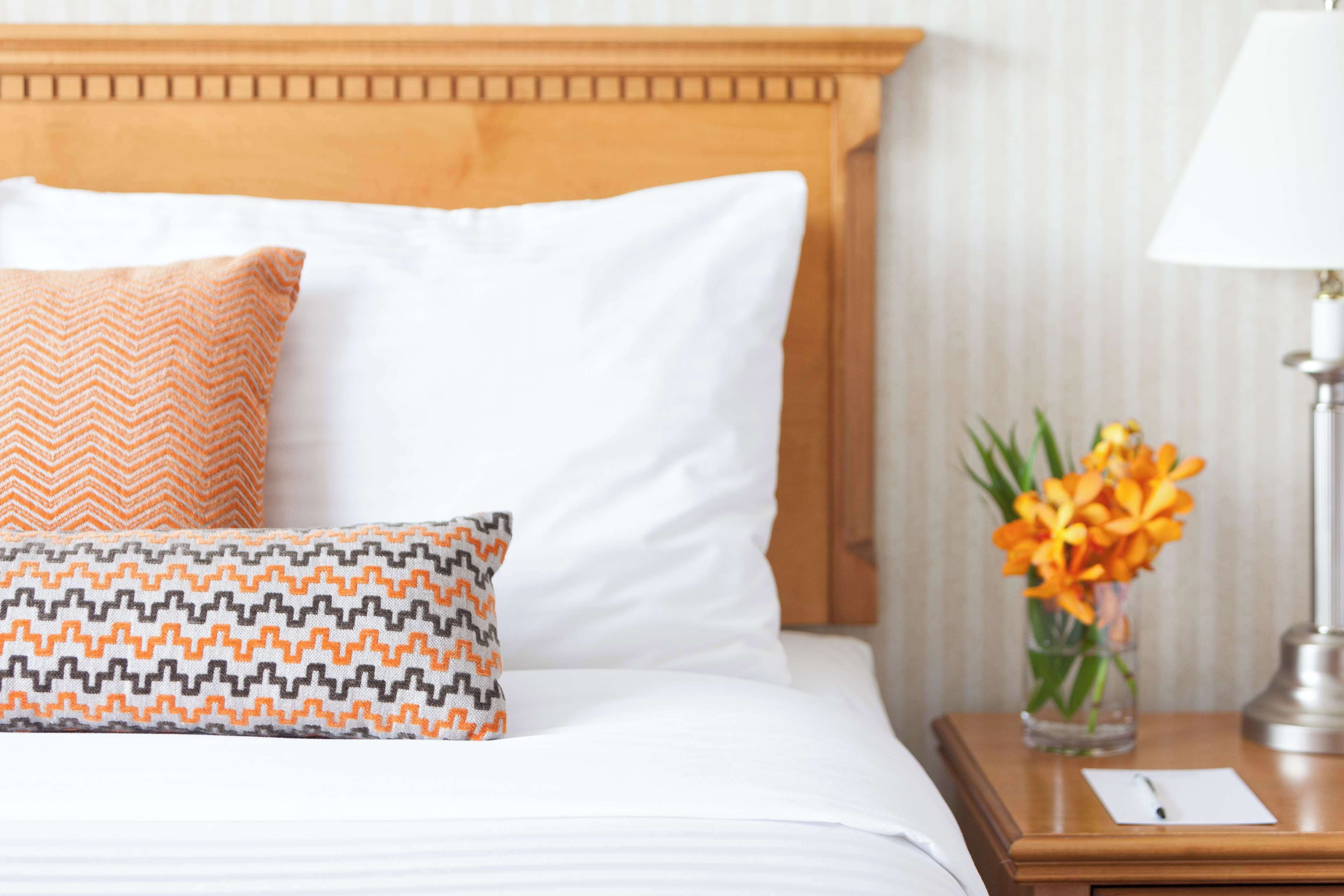 Una cama bien hecha con almohadas blancas y estampadas, un cabecero de madera, una mesita de noche con una lámpara y un florero con flores.