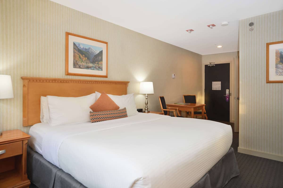 A hotel room with a large bed, a desk, and a chair. There is a painting on the wall and a door with a sign.
