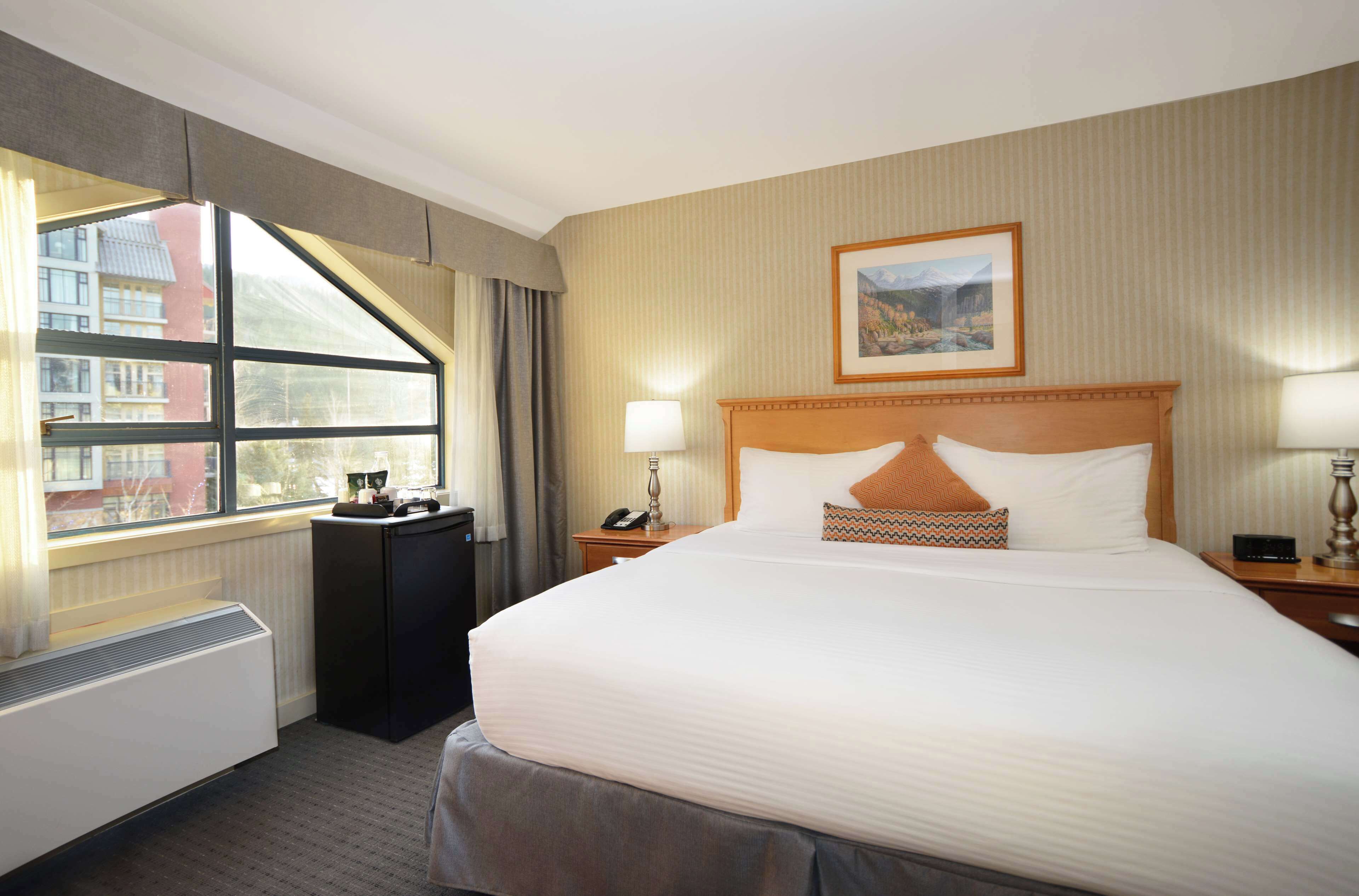 Hotelkamer met een groot bed, een raam met gordijnen, een radiator, een bureau met telefoon en een schilderij aan de muur.