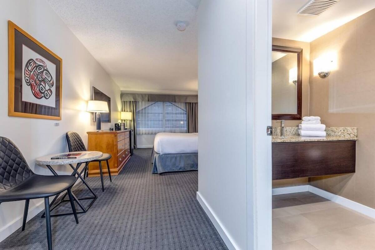 A hotel room with a bed, a desk with a chair, a small table, a dresser, and a bathroom with a sink and mirror.