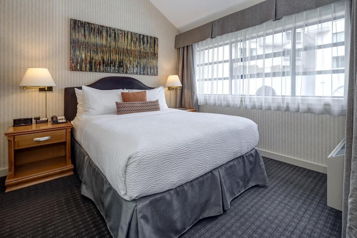 A hotel room with a large bed, two bedside tables with lamps, a modern abstract painting on the wall, and a large window with sheer curtains.