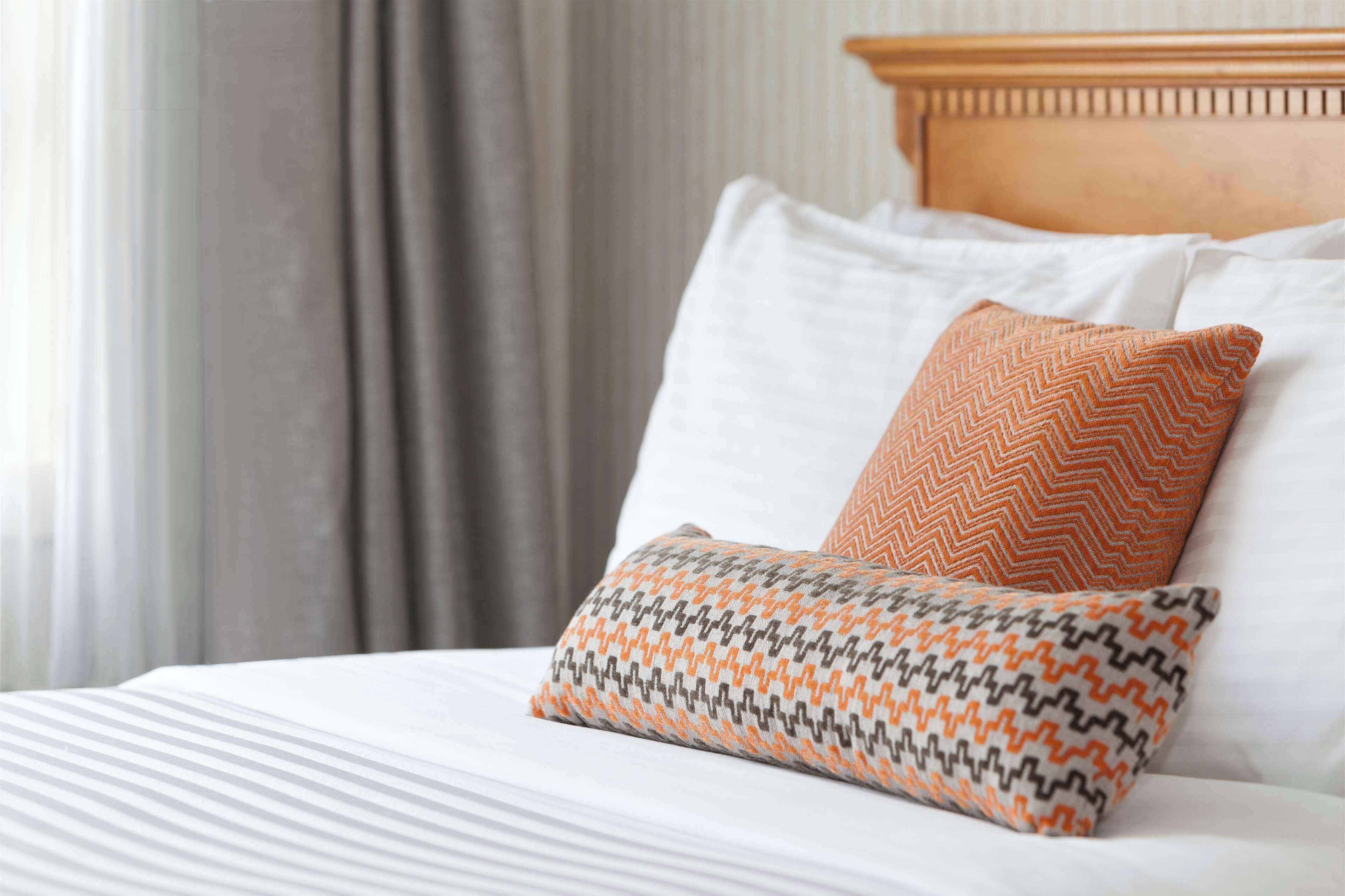 A neatly made bed with white linens and two patterned pillows, one orange and one gray and orange.