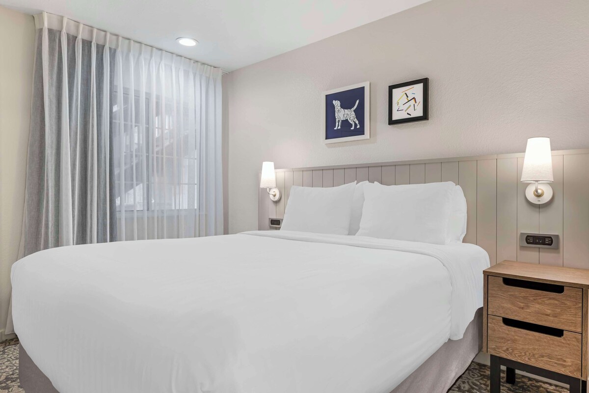 A hotel room with a large bed, two wall-mounted lamps, a nightstand with a lamp, and two framed pictures on the wall.